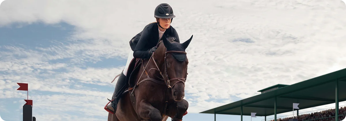 Un cavalier vêtu d'une tenue noire et d'un casque fait sauter un cheval brun par-dessus un obstacle rayé rouge et blanc lors d'un concours de saut d'obstacles devant les gradins d'un stade.